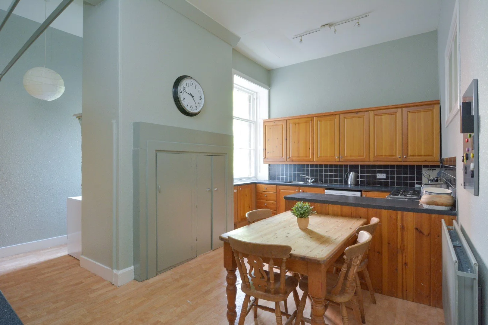 Kitchen with wooden cabinets, black countertop, small dining table with four chairs, potted plant, window, and wall clock.