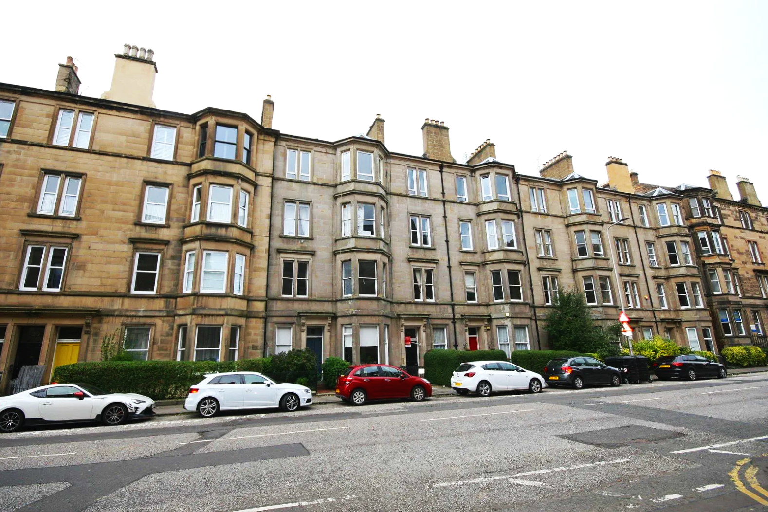 A corner view of a sandstone multi-story apartment building with bay windows, on a city street with parked cars and a blue sky with clouds.