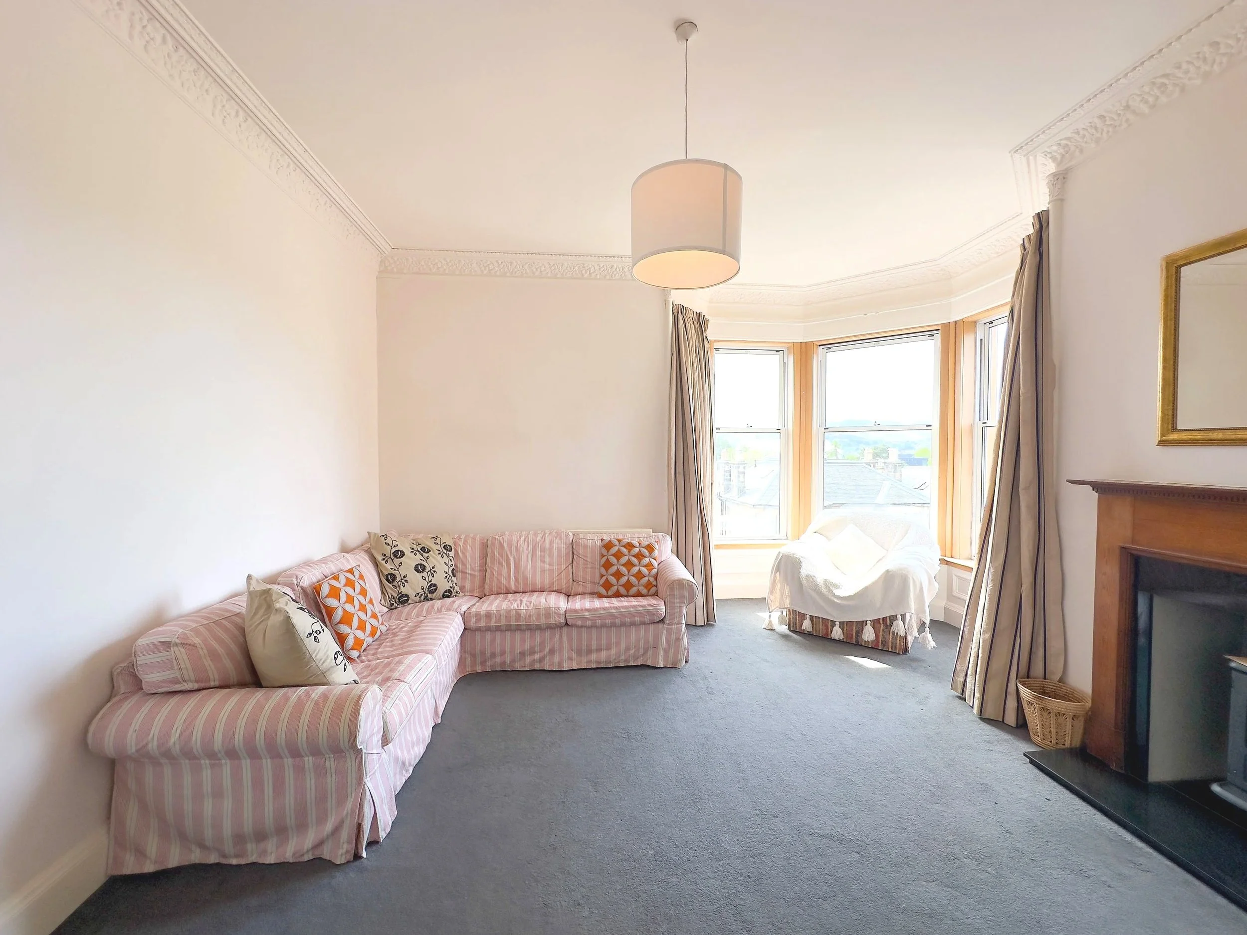 Living room with a pink striped sofa, patterned pillows, a white armchair by large bay windows, and a fireplace with a mirror above it.