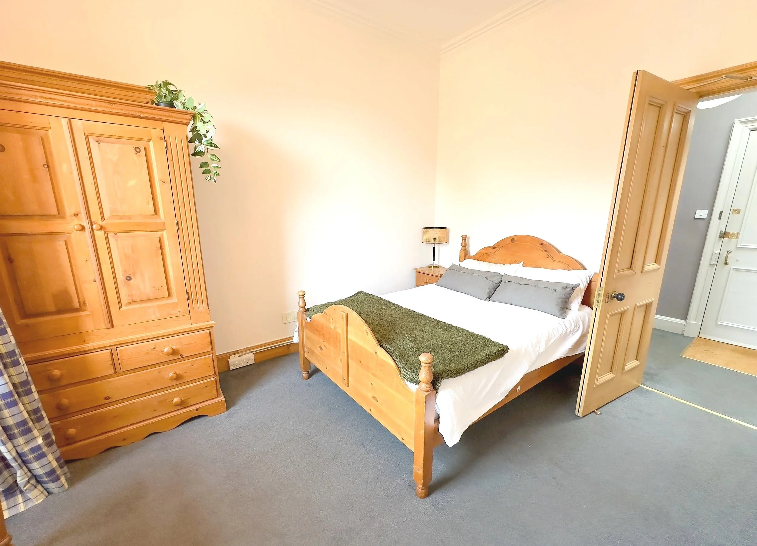 A bedroom with a wooden bed frame, white bedding, gray pillows, a green throw blanket, a wooden wardrobe, a bedside table with a lamp, and a door open to a gray wall hallway.