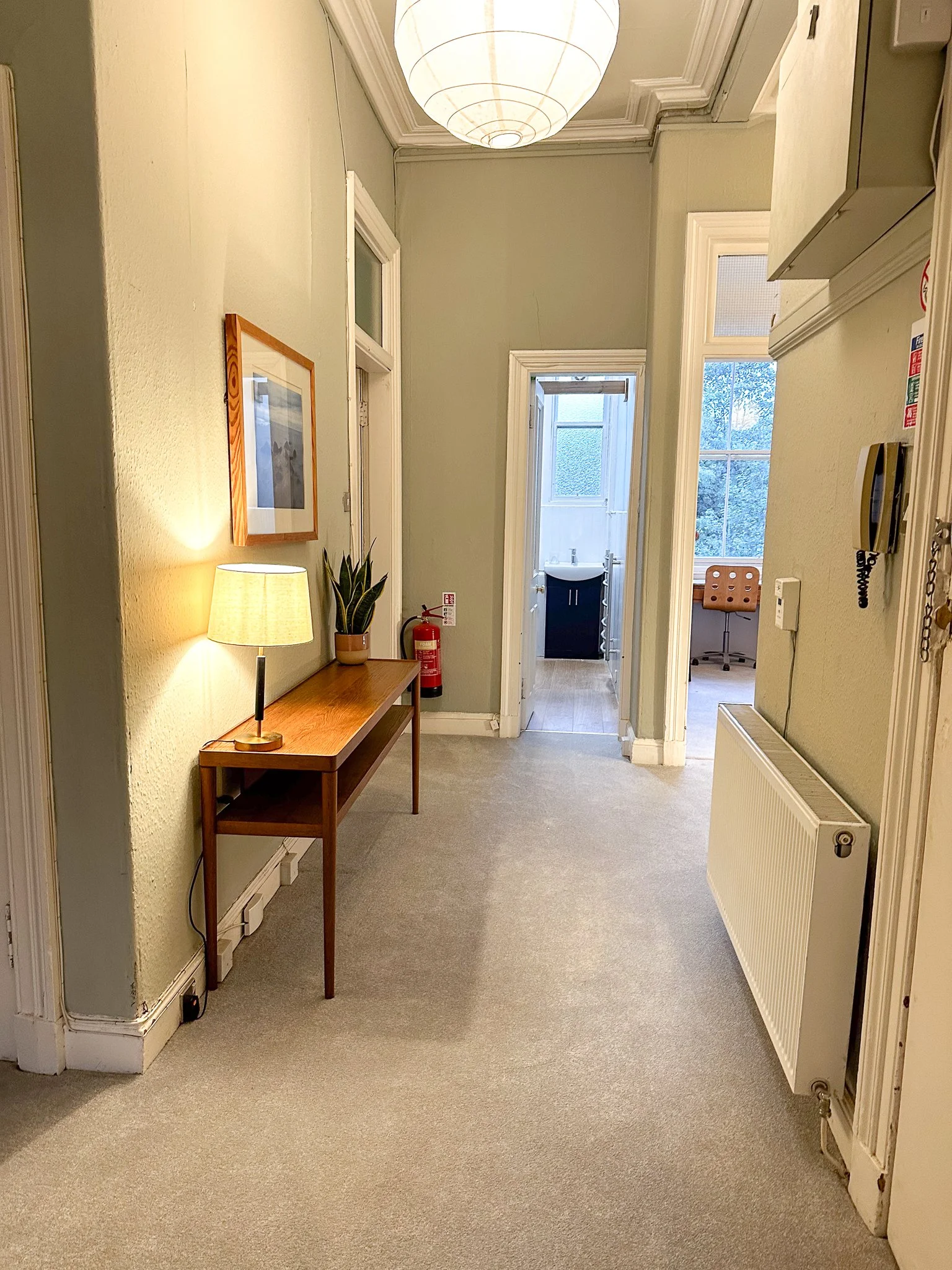 A hallway with a wooden table, a lamp, a fire extinguisher, and a potted plant on the table. The hallway has light-colored walls, a radiator, and multiple doors leading to rooms with windows and a sink visible. A large round paper lantern hangs from the ceiling.