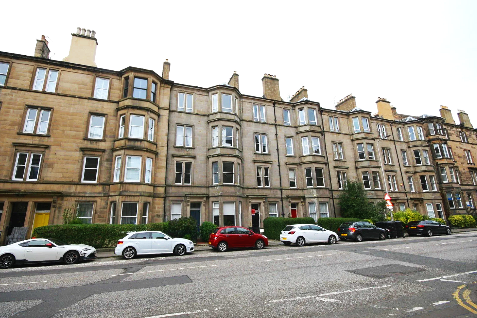 Street view of a multi-story stone tenement building with multiple bay windows, green bushes, and parked cars along the curb.