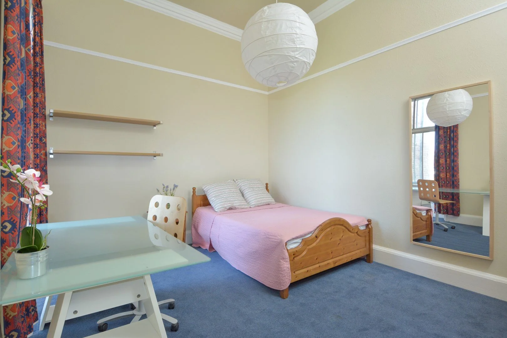 A bedroom with a wooden bed frame, pink bedding, and striped pillows, a glass table with a potted orchid, a large wall mirror, a window with red patterned curtains, and a white paper lantern ceiling light.