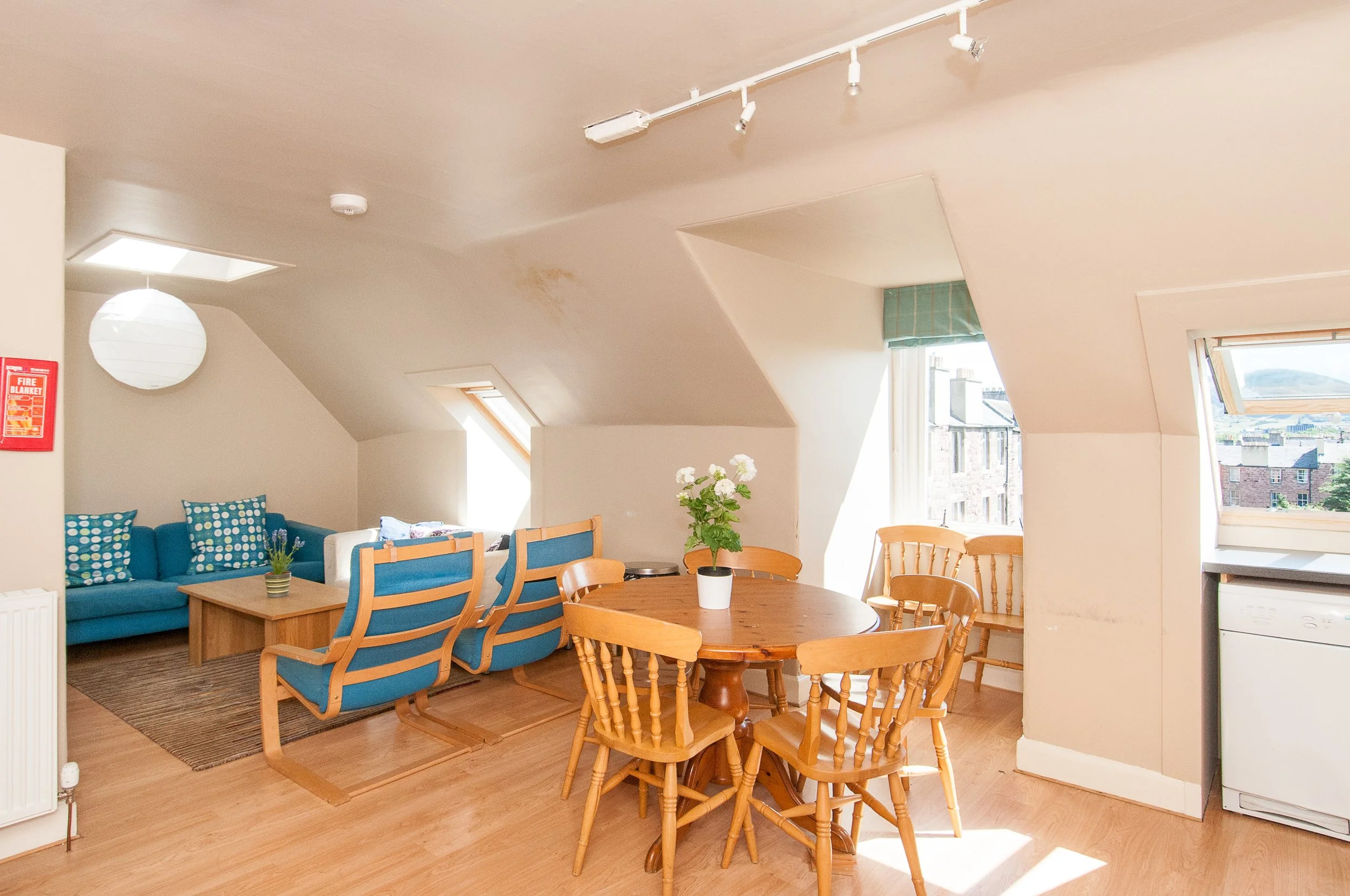 Modern wooden house on a grassy landscape with small trees near a lake, with mountains in the background under a cloudy sky.