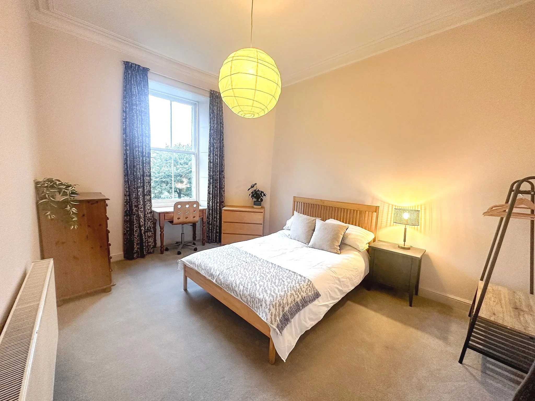 A bedroom with a bed, nightstand, table lamp, dresser, small desk with a chair, window with curtains, and a clothing rack.