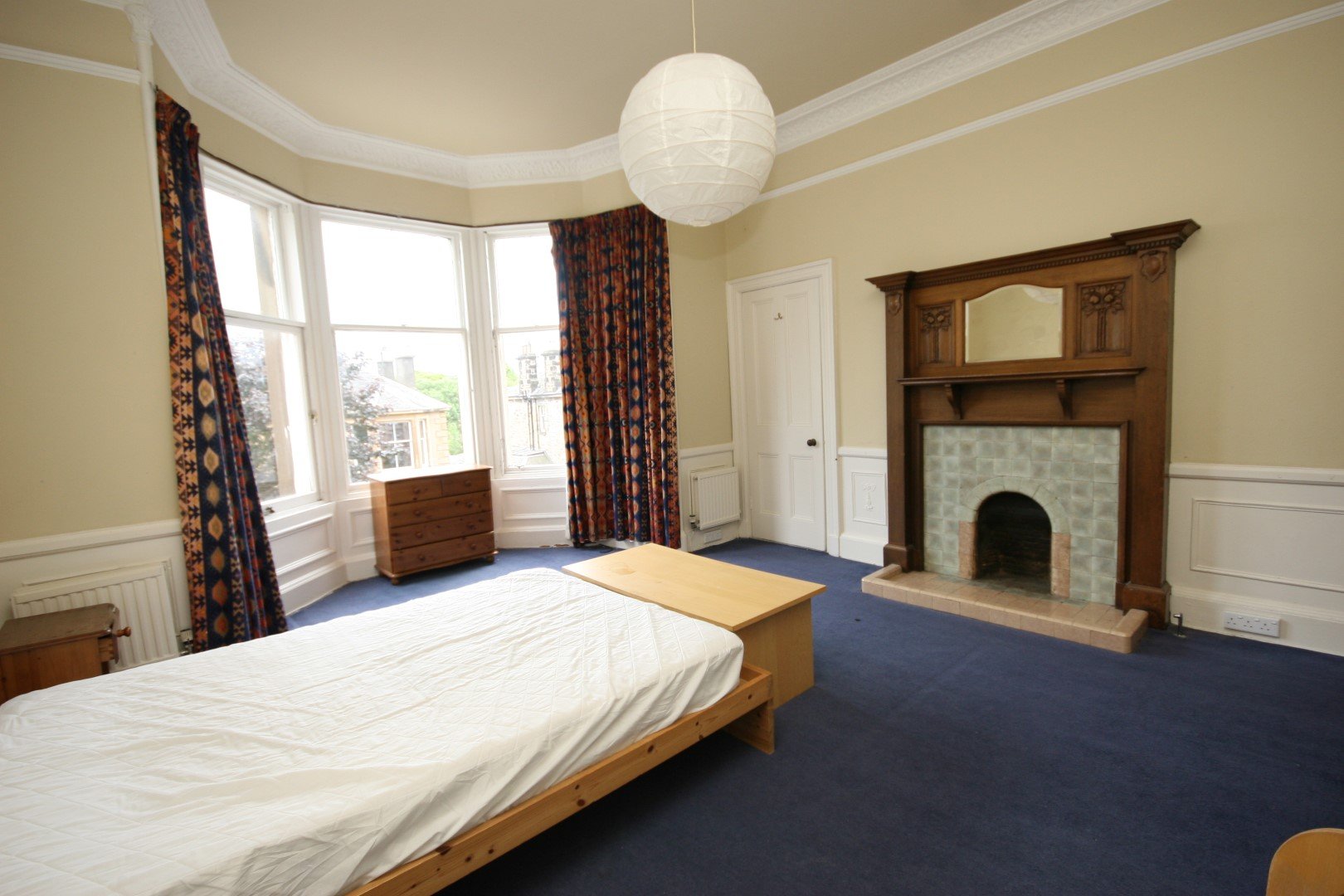 A bedroom with a bed, a wooden dresser, a fireplace with intricate wooden mantel, large bay window with colorful curtains, dark blue carpet, and cream-colored walls with white trim.