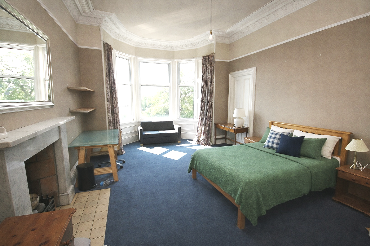 A cozy bedroom with a large bay window, a green bedspread on a wooden bed, a small sofa near the window, a fireplace, and side tables with lamps.