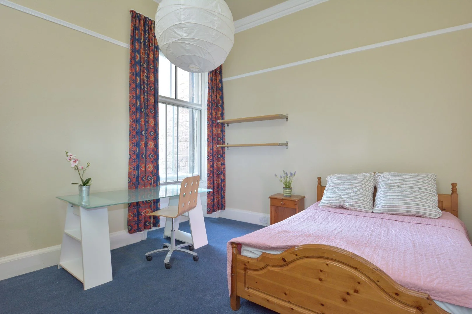 A cozy bedroom with a wooden bed frame, pink quilt, two striped pillows, two floating shelves, a nightstand with a plant, a glass desk with a flower and a chair, a large window with colorful curtains, and a paper lantern ceiling light.