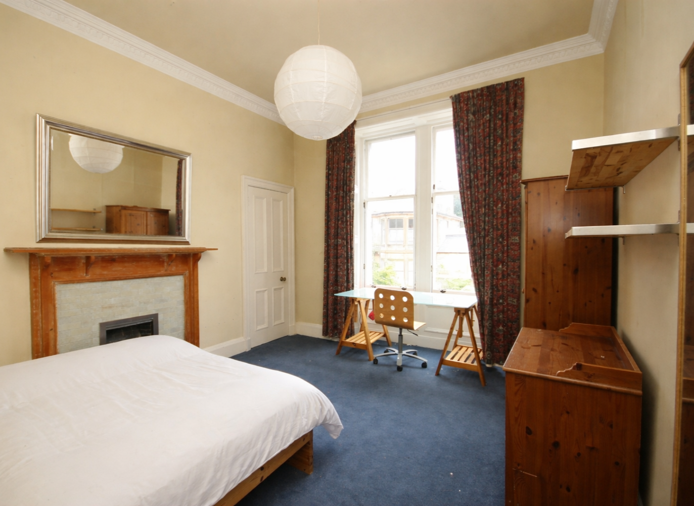 Bedroom with a bed, fireplace with a mirror above, wooden shelves and furniture, a desk near large window with curtains, and a paper lantern ceiling light.