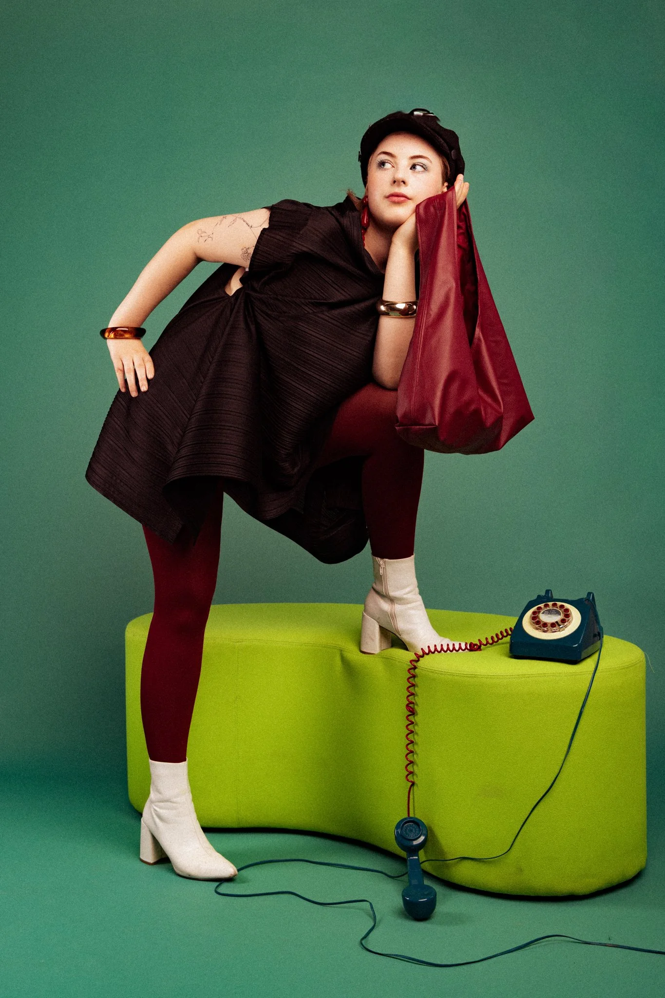 A woman with short dark hair wearing a black top, maroon tights, white boots, a black hat, and gold jewelry leans on a red bag, standing on a green curvy bench in front of a green background. There is a vintage rotary phone with a red cord and a blue