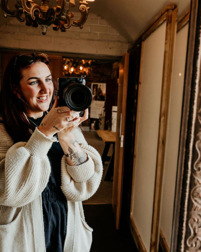 Woman taking a selfie with a camera in a cozy, warmly lit interior with rustic décor, chandelier, and mirror.