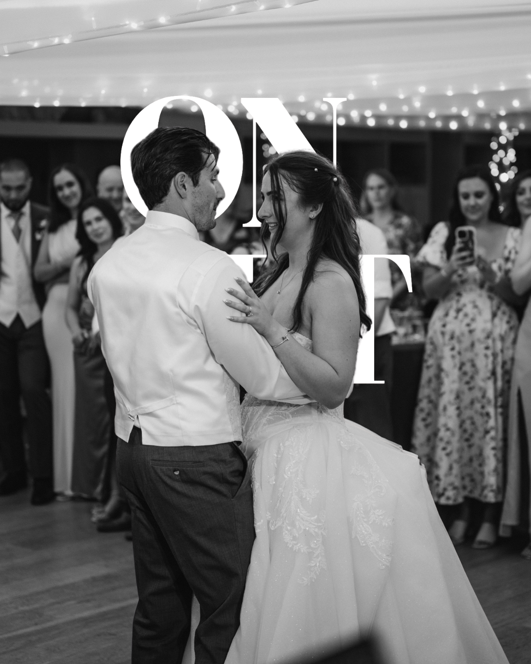 A black and white photo of a bride and groom dancing at their wedding reception, surrounded by guests who are watching and taking photos.