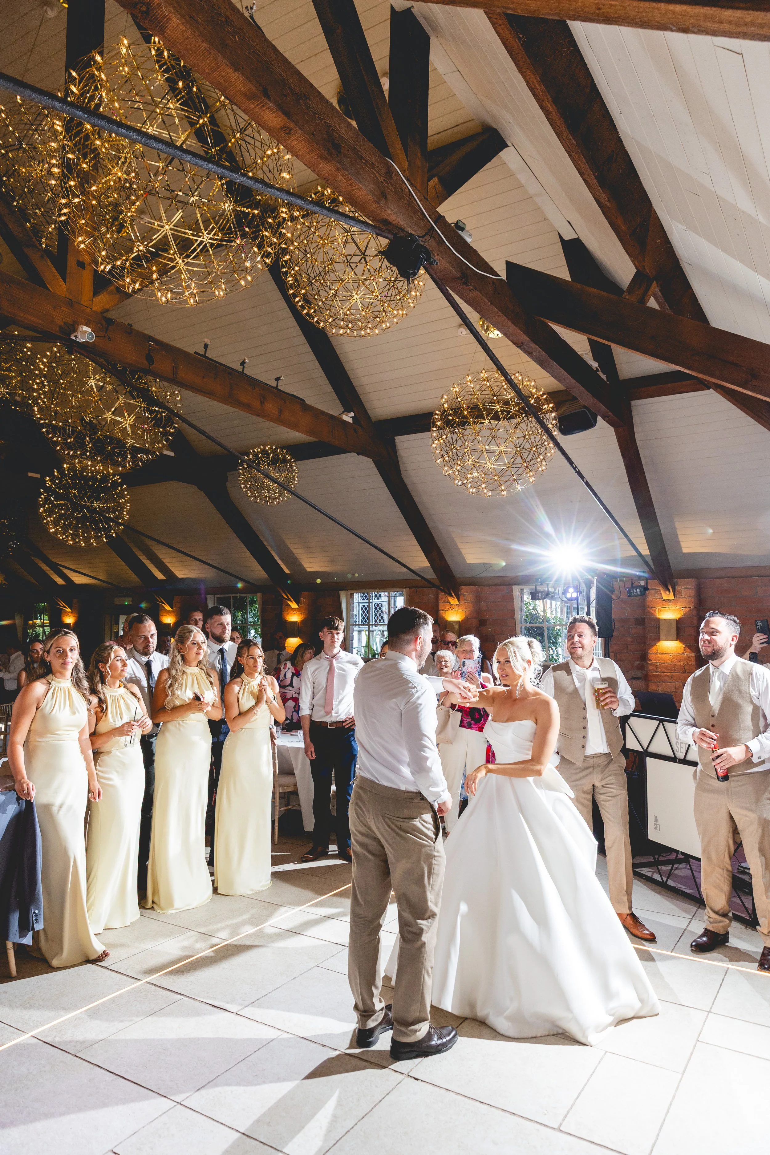 Wedding reception scene with a bride and groom dancing, surrounded by friends and family in a decorated venue with golden chandeliers.