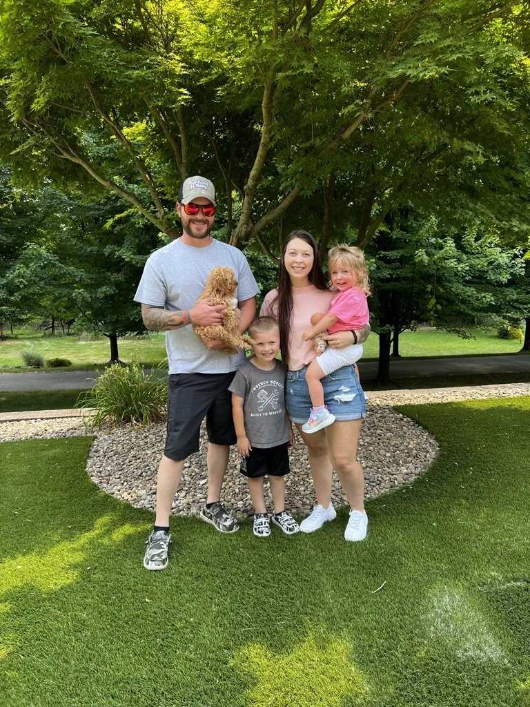 Family of four with two children and a dog posing outdoors on a grassy lawn with trees in the background.