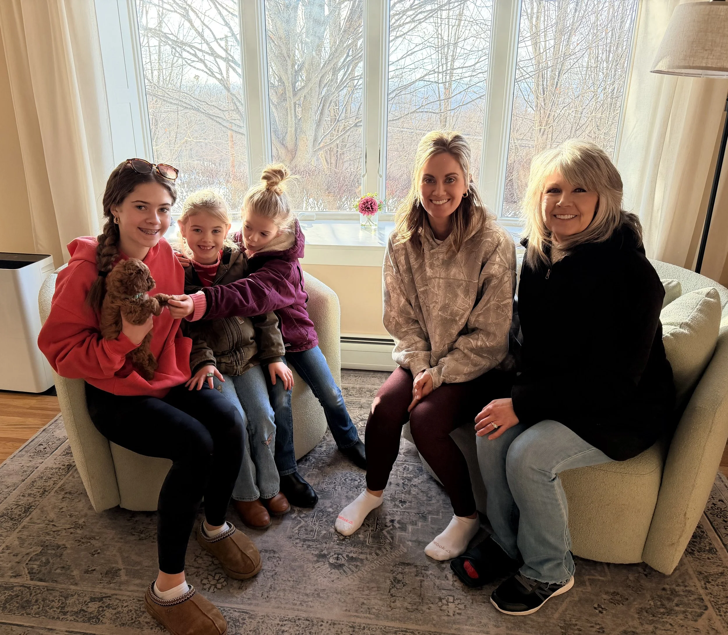Group of five women and children sitting on two sofas in a living room, with large windows overlooking a winter landscape, and a small pink flower in a vase on the window sill.