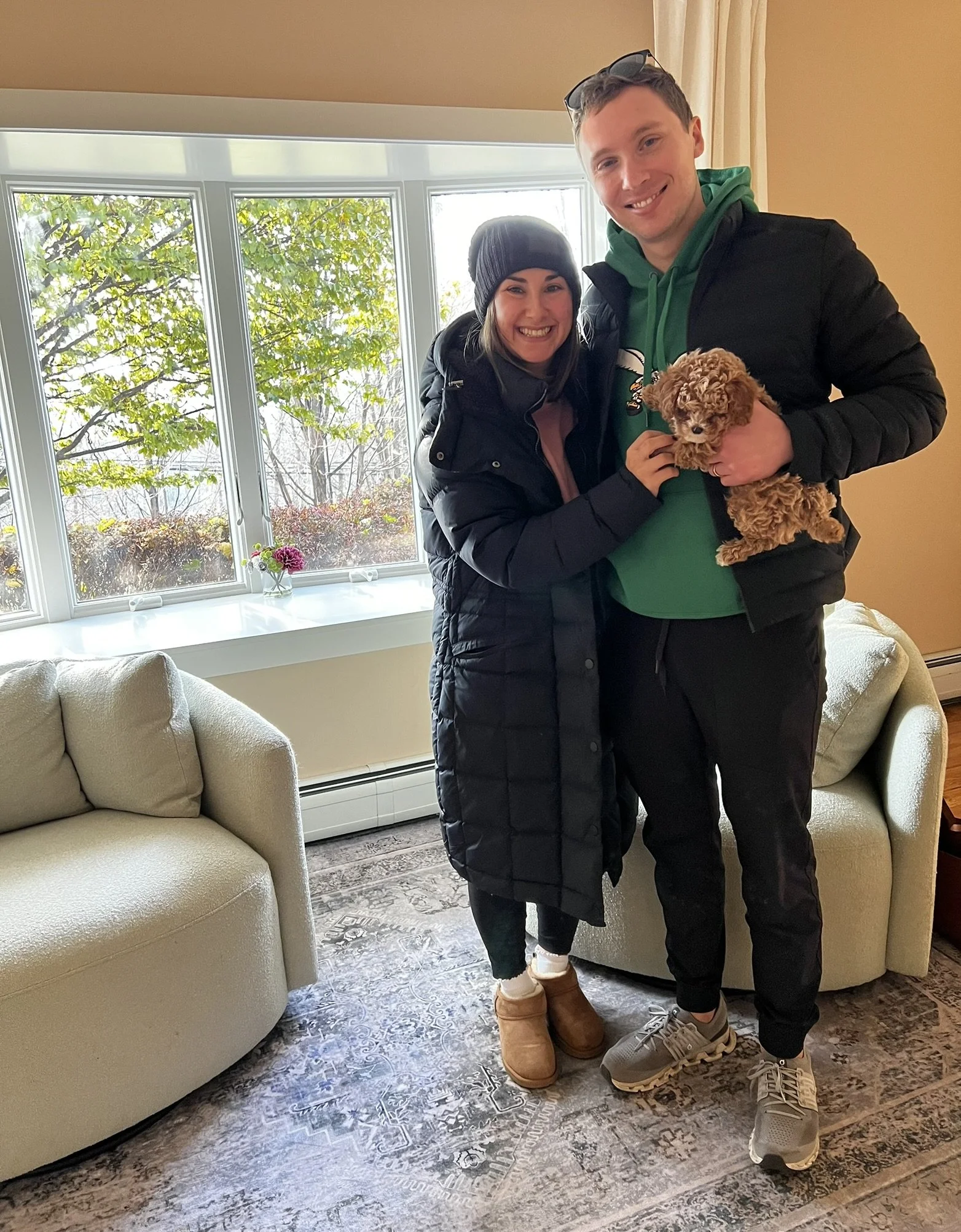 A smiling couple holding a small brown poodle puppy, standing inside a room with large window, beige walls, and cream-colored furniture.