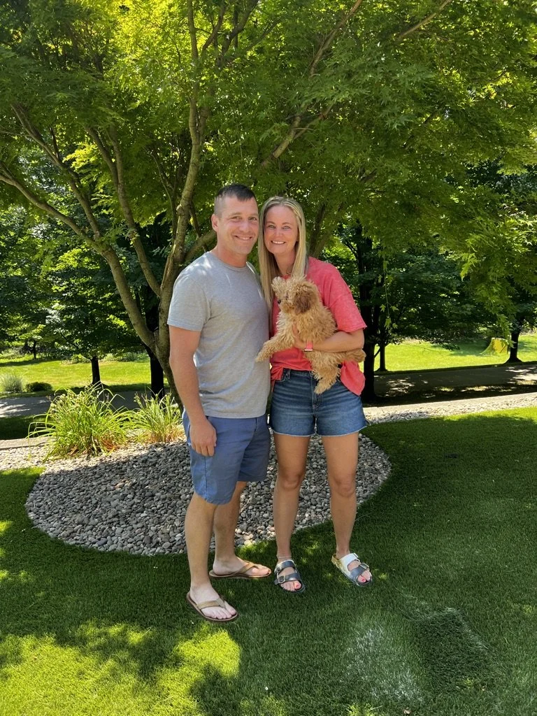 A smiling couple standing under a large green tree on a sunny day, with the woman holding a fluffy brown dog, in a park setting with grass and small plants.