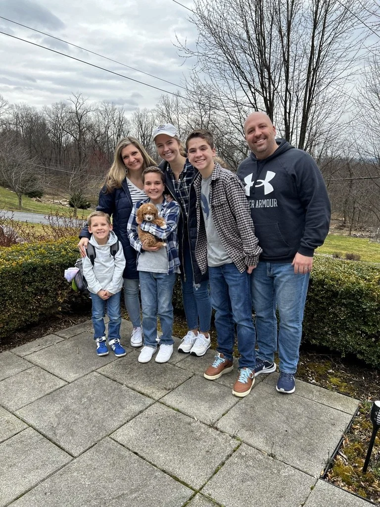 Family of seven standing on concrete patio outdoors with trees and bushes in the background, cloudy sky overhead. They are smiling, with one child holding a small brown puppy.