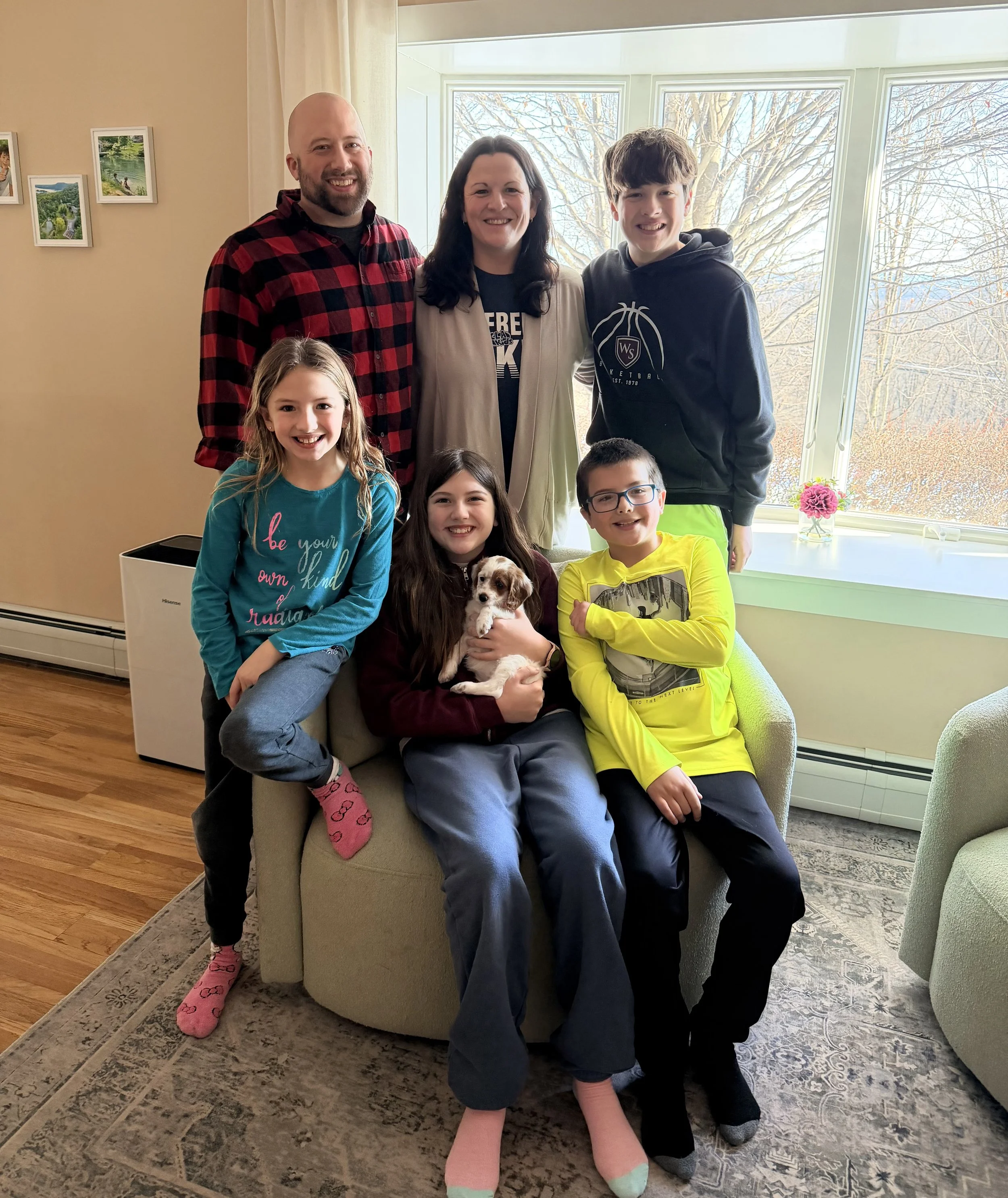 A family photo inside a living room shows three adults and four children, with one holding a puppy, standing in front of a large window with a view of trees and mountains outside.