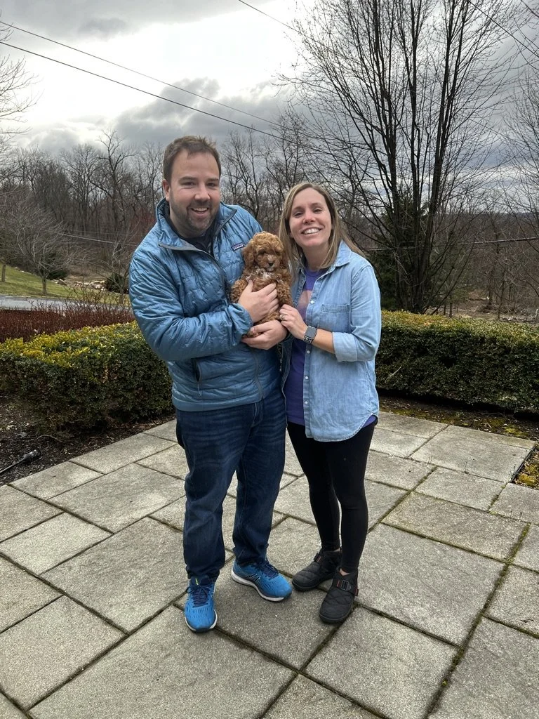 A man and woman standing together outdoors on a cloudy day, smiling and holding a small brown puppy between them, with leafless trees and bushes in the background.