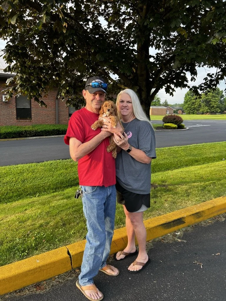 A man and woman standing outdoors on a parking lot curb, holding a small brown puppy between them, with a large tree and a brick building in the background.