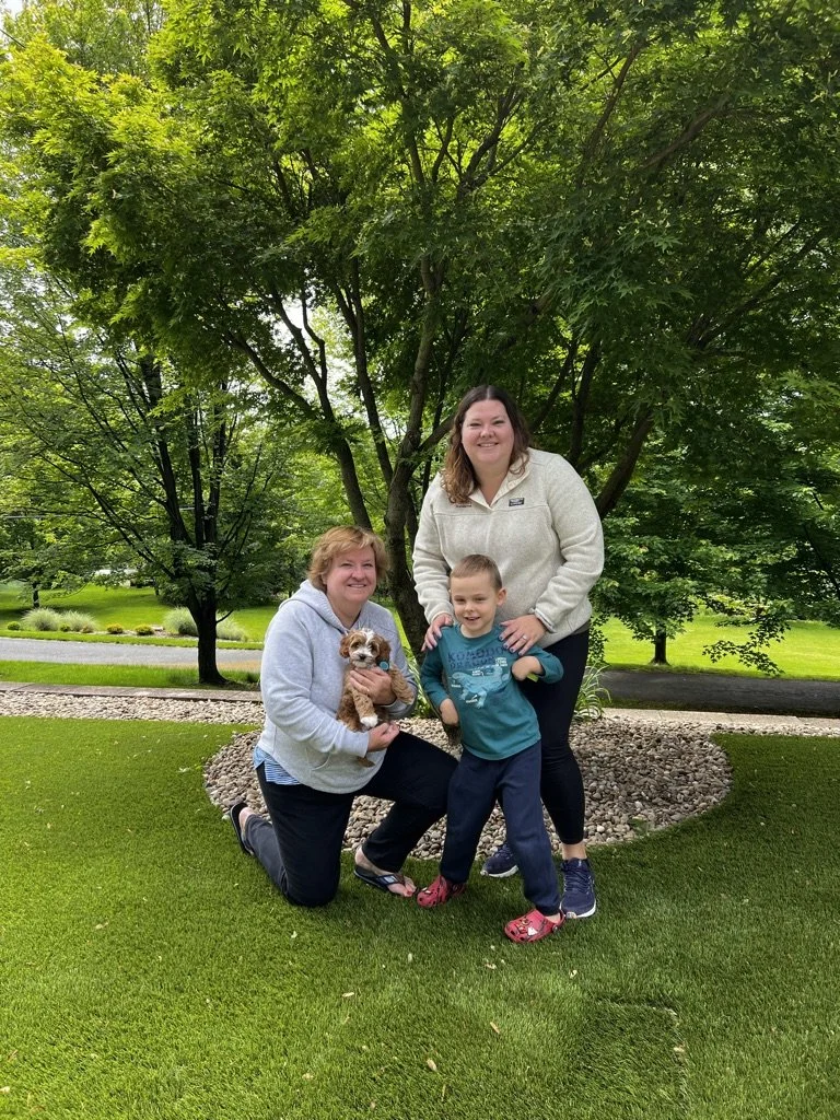 Family of three with a small dog enjoying time outdoors in front of large green trees and a neatly landscaped yard.