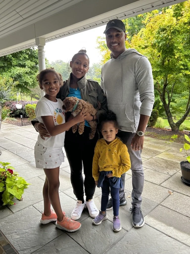 A family of five, including two women, a man, and two young girls, are standing together on a porch with trees and greenery in the background. One woman is holding a small brown puppy.