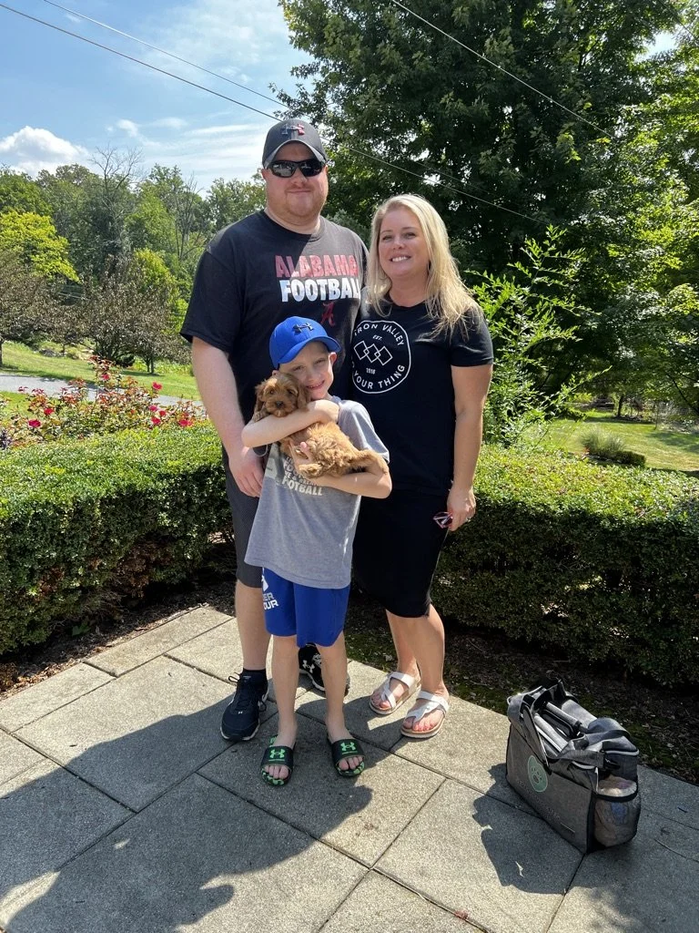 A smiling family of three with a dog outdoors on a sunny day, standing on a paved walkway with greenery and trees in the background.