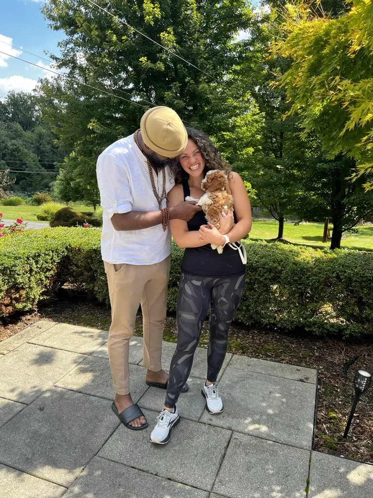 A man and woman are standing outside on a paved path, smiling and holding a small brown and white puppy. The man is wearing a tan beanie, white shirt, beige pants, and sandals, while the woman is dressed in black workout clothes with patterned leggin