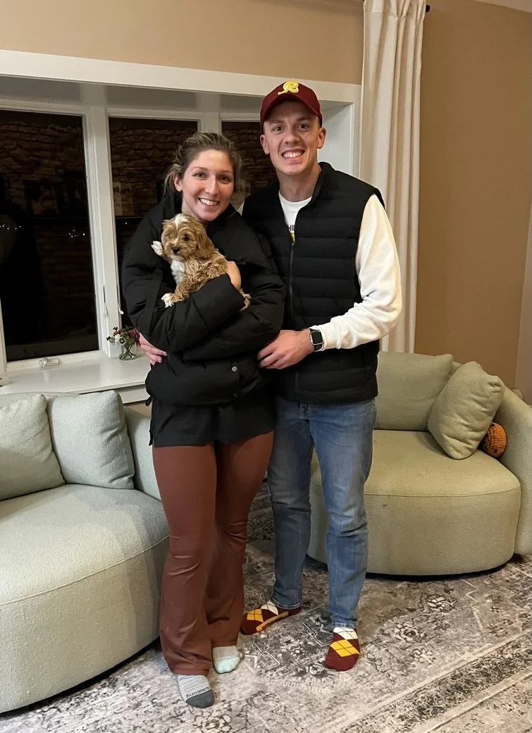 A smiling woman and man stand together in a living room, holding a small dog. The woman is wearing a black jacket and brown pants, while the man is wearing a white sweatshirt, a black vest, and colorful socks. There is a beige sofa and a window behin