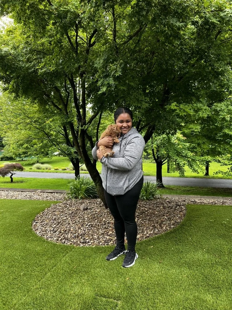 A woman holding a small curly-haired dog standing on a grassy lawn with trees and a rock bed in the background.