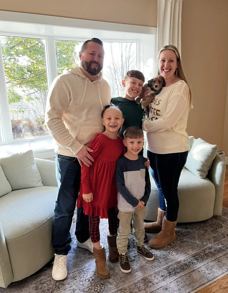 A family of five, including two adults and three children, poses together indoors near a large window with a dog. The family is smiling, and the kids are dressed in casual clothes, with one girl in a red dress and two boys in striped and solid shirts