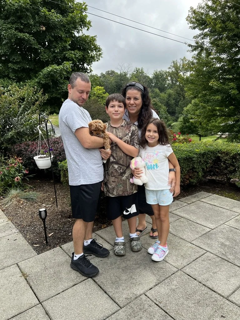 A family consisting of a man, a woman, a boy, and a girl standing outdoors on a stone patio. The man is holding a small brown puppy, and the girl is holding a plush toy. The background features greenery, trees, and bushes under a cloudy sky.
