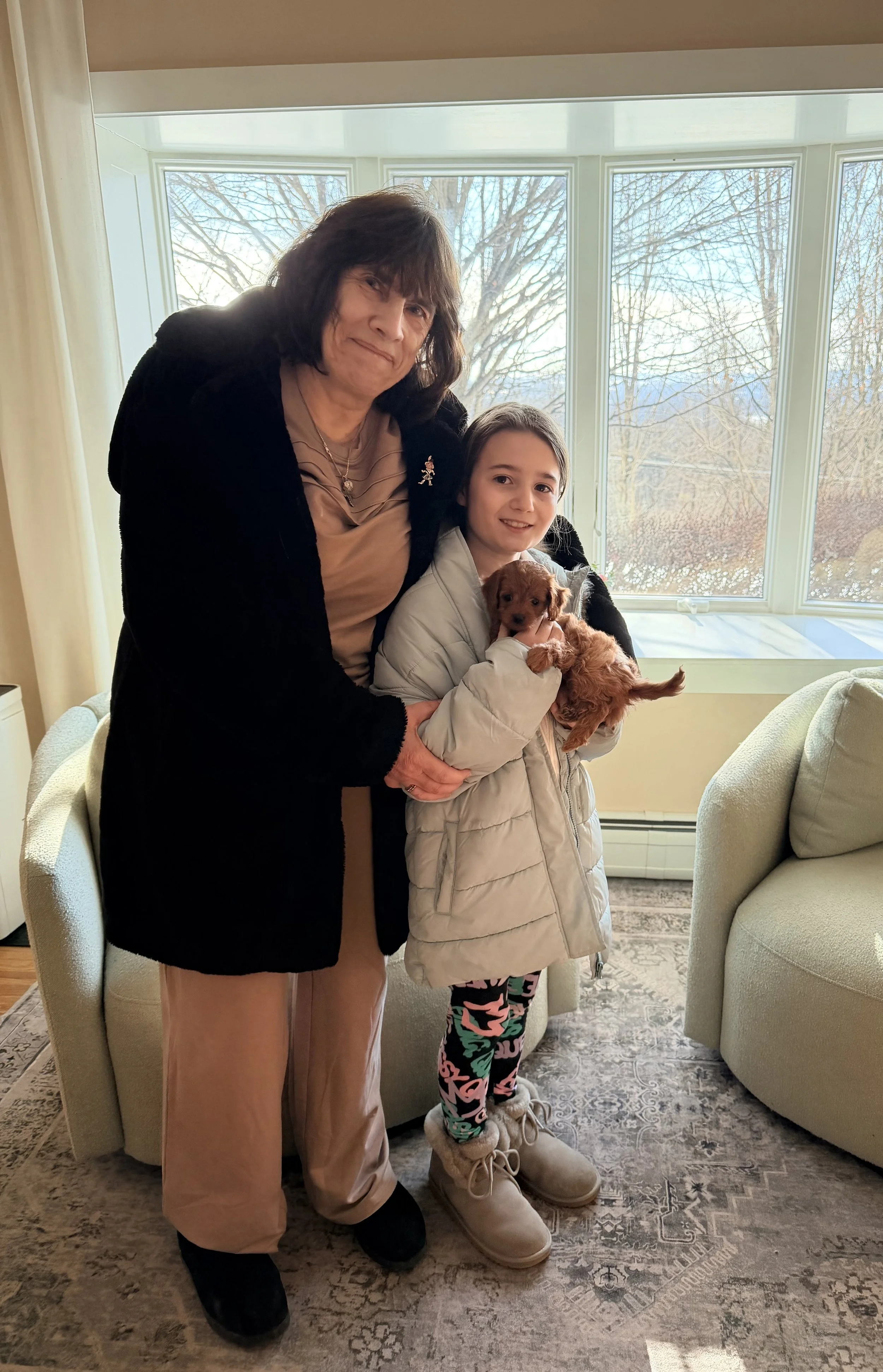 A woman and a girl are standing indoors by a large window, holding a small brown puppy. The woman has dark hair and is wearing a black coat, beige pants, and black shoes. The girl has shoulder-length hair and is wearing a light-colored coat, colorful