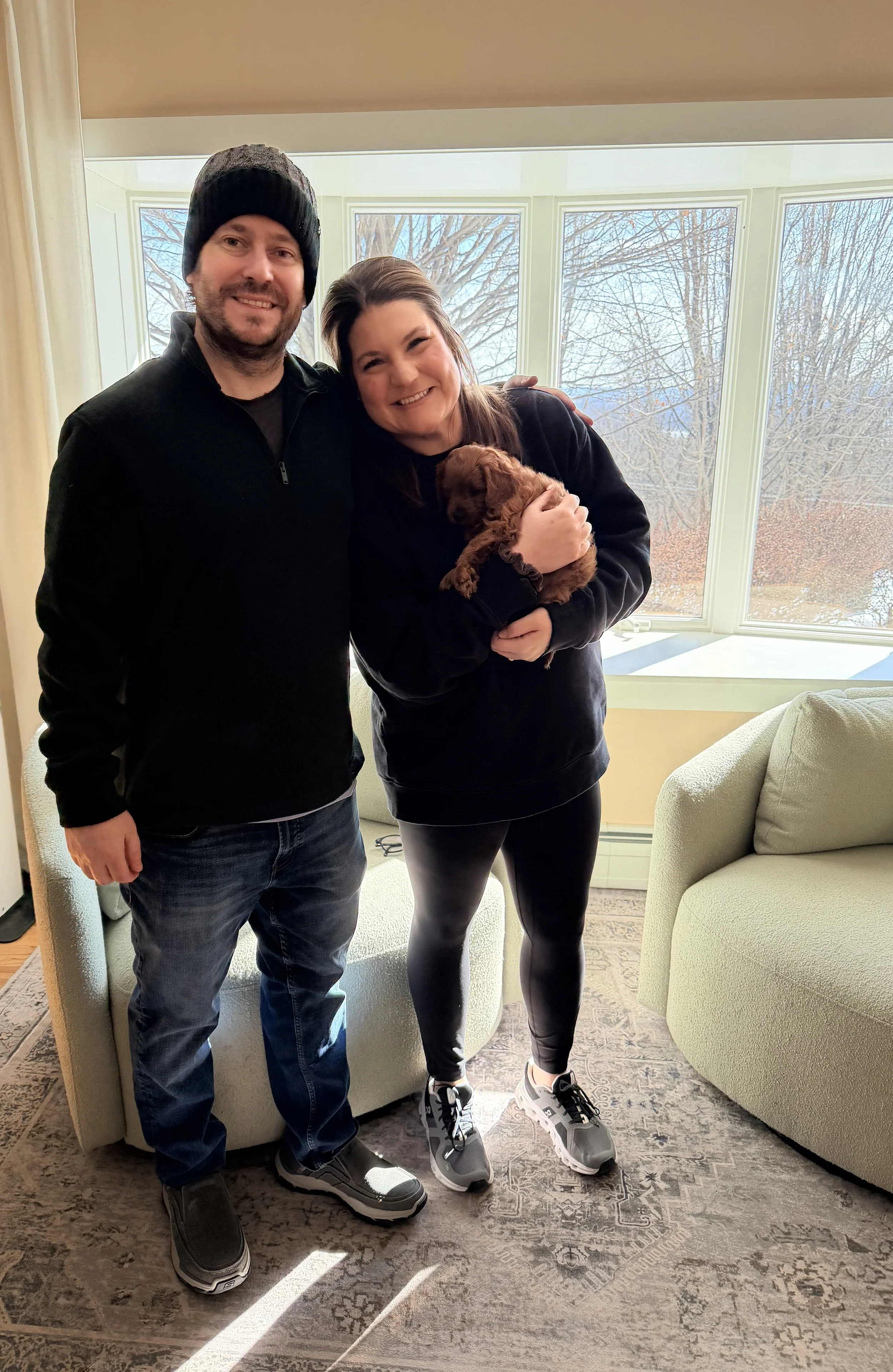 A man and woman standing together indoors, smiling, holding a small brown puppy, with large windows showing a winter landscape outside.