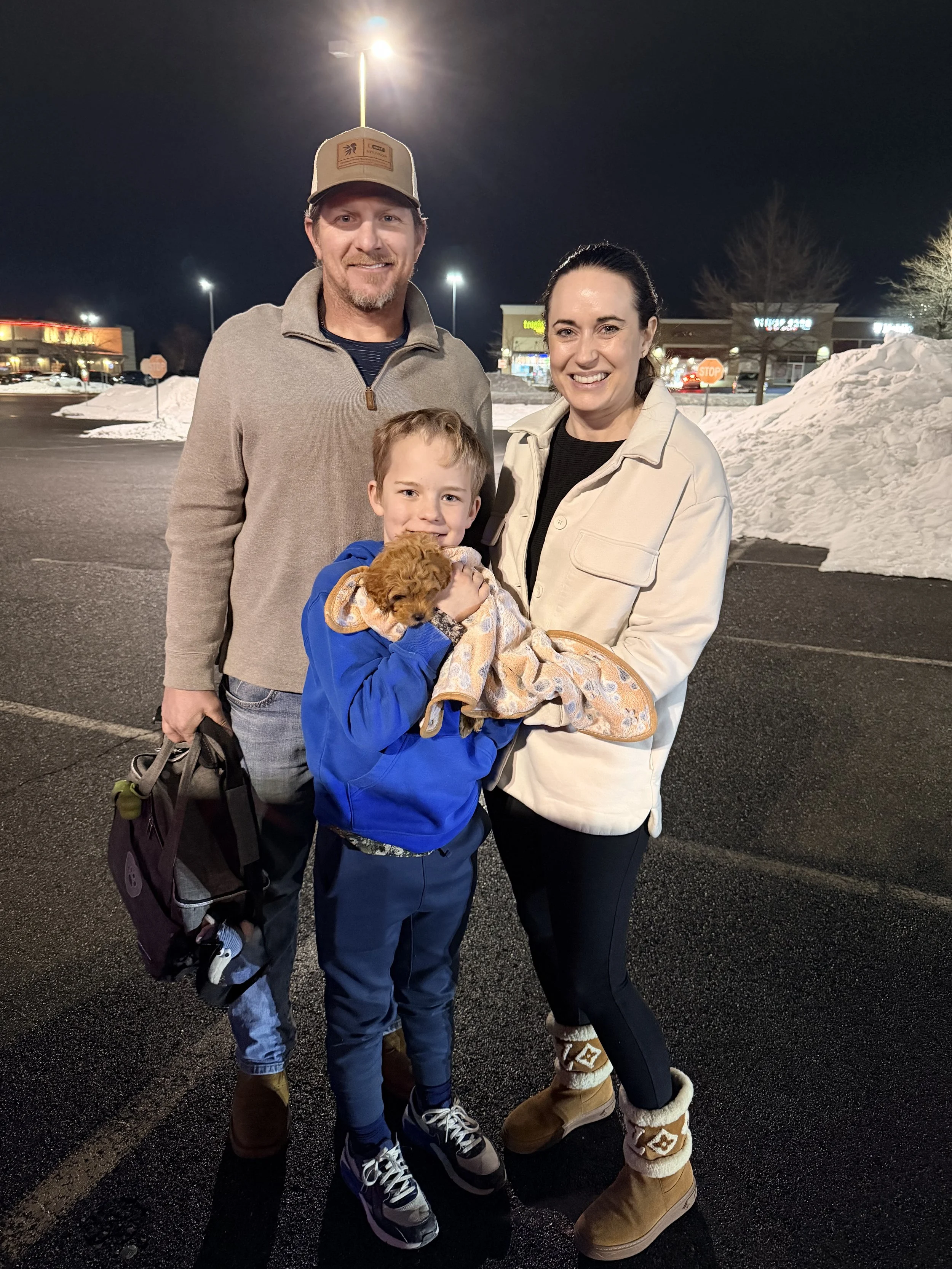 Family of four stands in a parking lot at night, smiling. The father is wearing a beige cap and gray sweater, the mother in a beige jacket, a boy in a blue hoodie holding a small brown puppy. Snow piles are visible in the background.