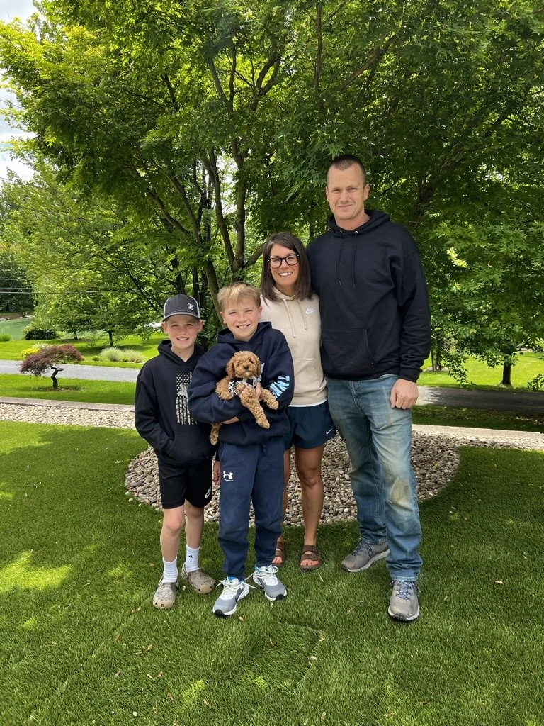 A family of four with a small dog standing on a well-manicured lawn in front of a large green tree on a sunny day.