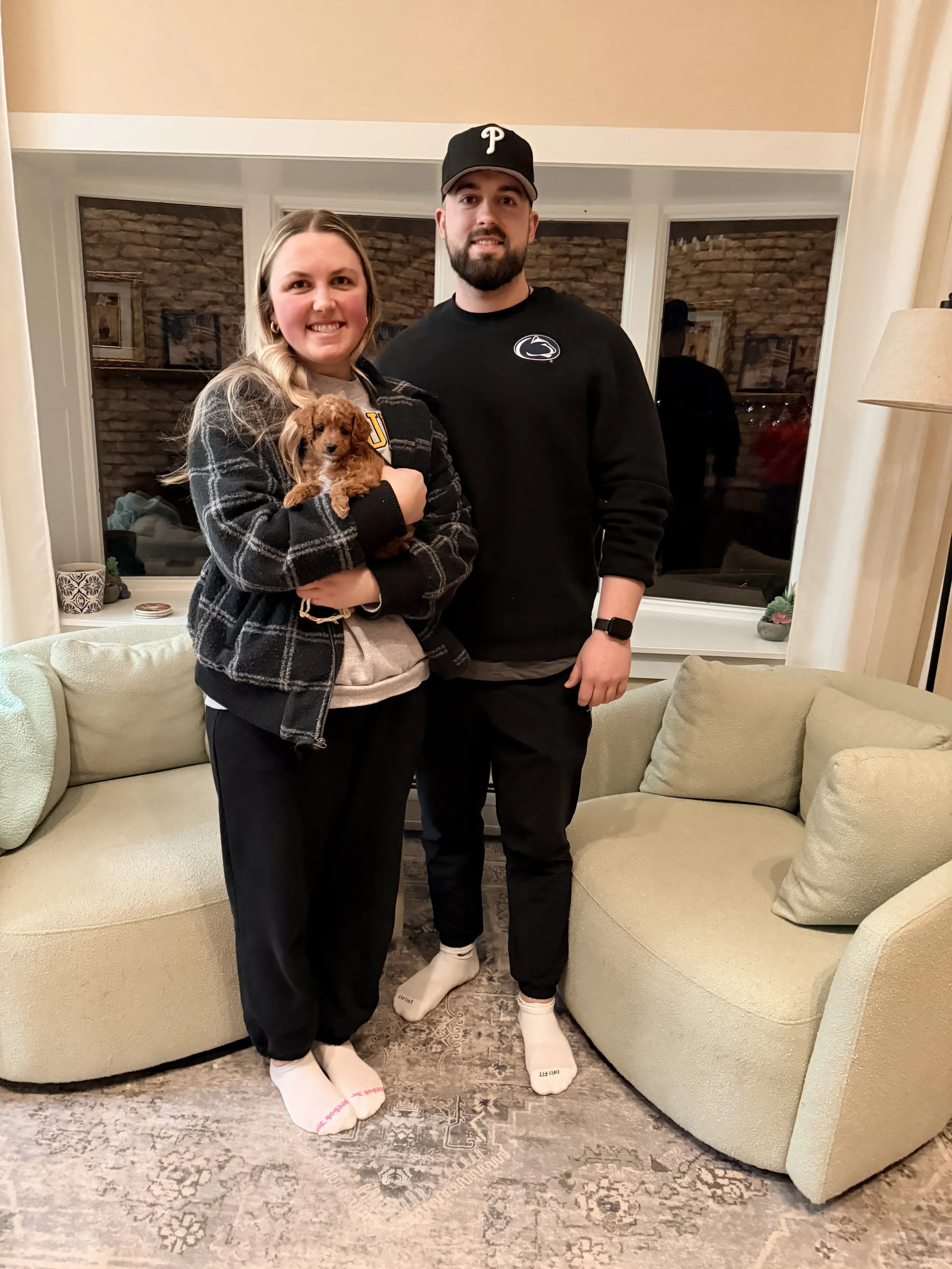 A smiling woman holding a small brown puppy next to a tall man in a black Penn State sweatshirt, all standing in a living room with beige furniture and a window in the background.