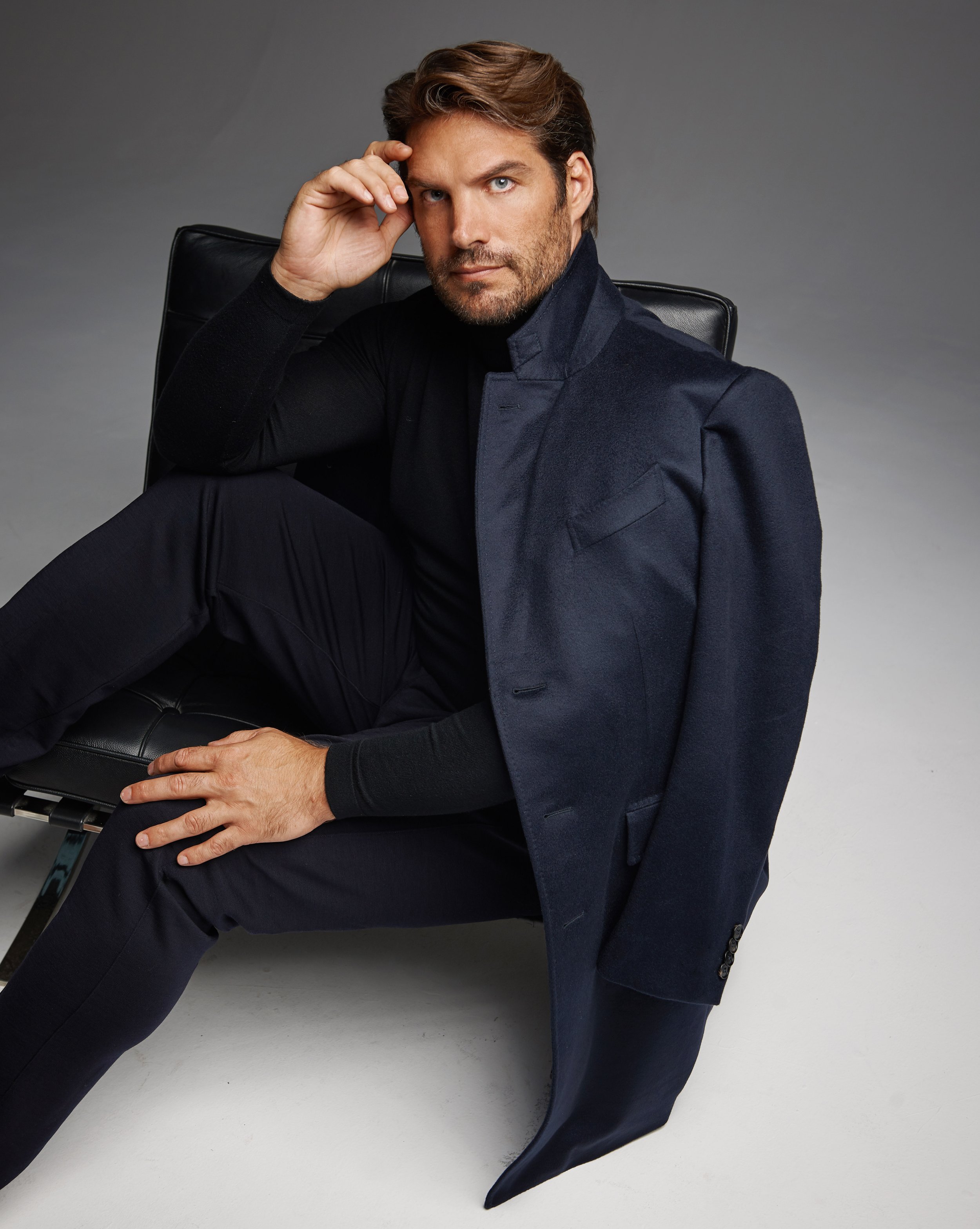 Homme en costume sombre assis sur une chaise noire, regardant vers l'avant, dans un studio avec fond gris.