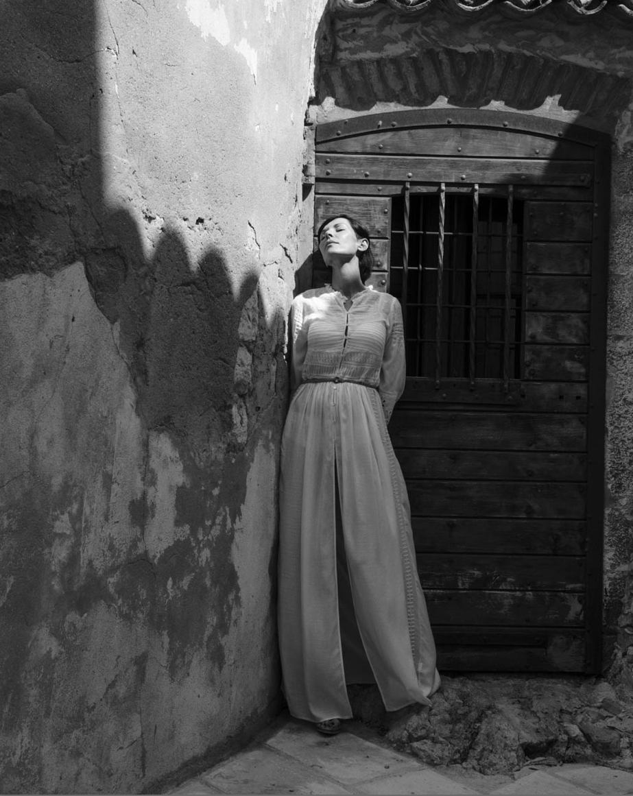 Femme debout contre un mur en pierre, yeux fermés, dans une cour avec une porte en bois, en noir et blanc.