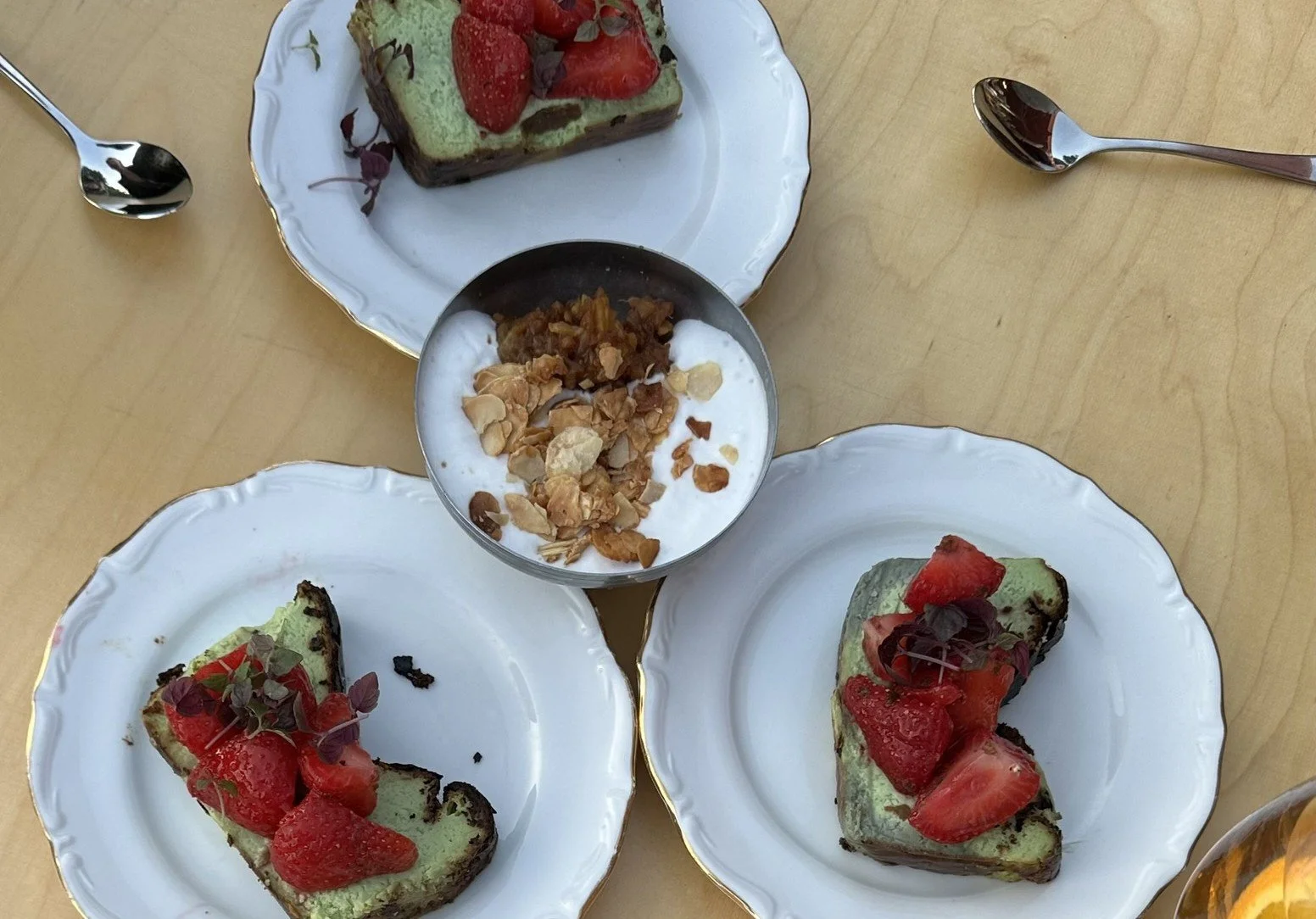 Three white plates with slices of green cake topped with strawberries and microgreens, a bowl of yogurt with granola and nuts, and a dessert bar with strawberries and microgreens, all on a wooden table.
