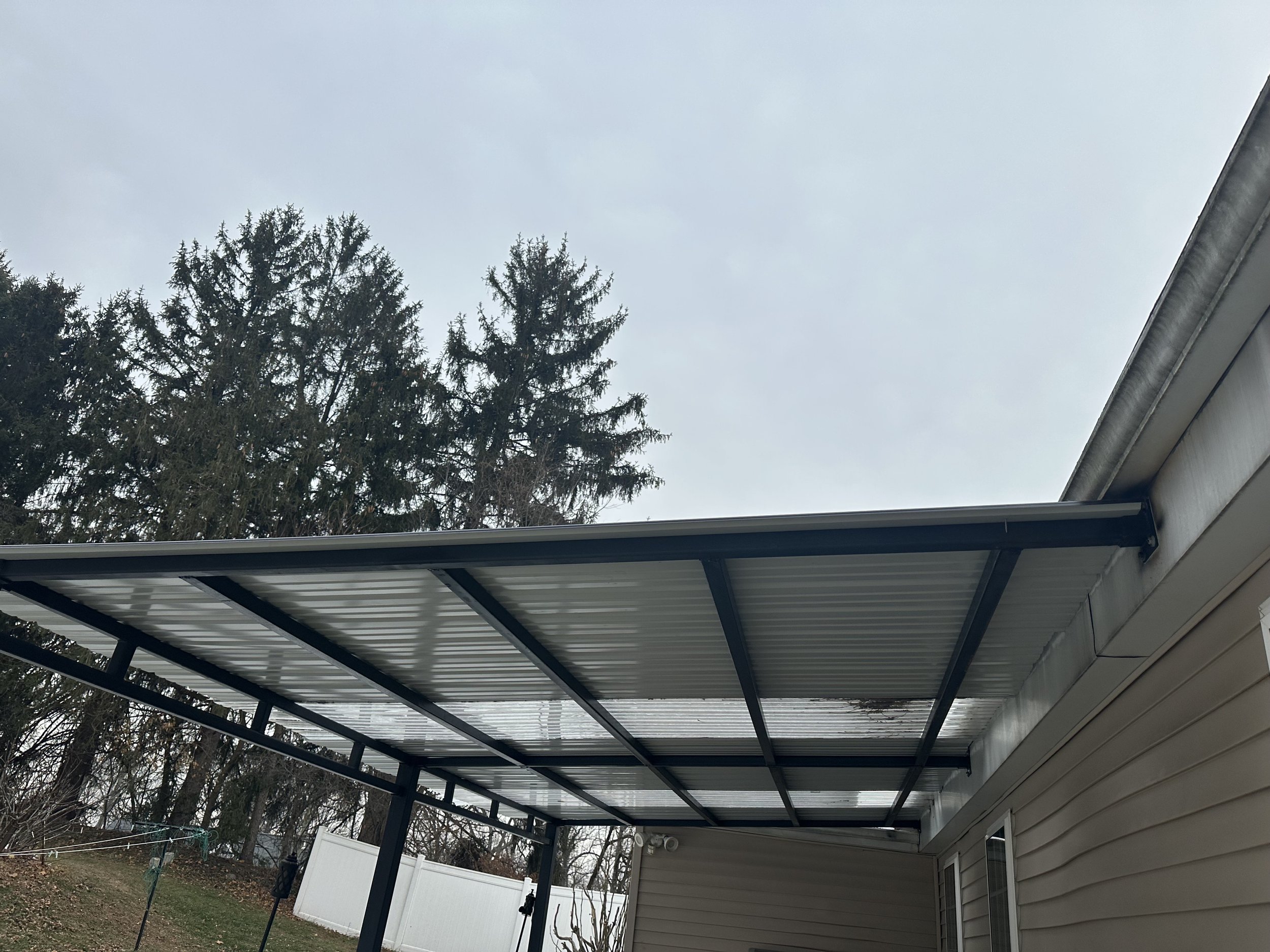 Metal carport attached to a beige house with trees in the background and a cloudy sky.