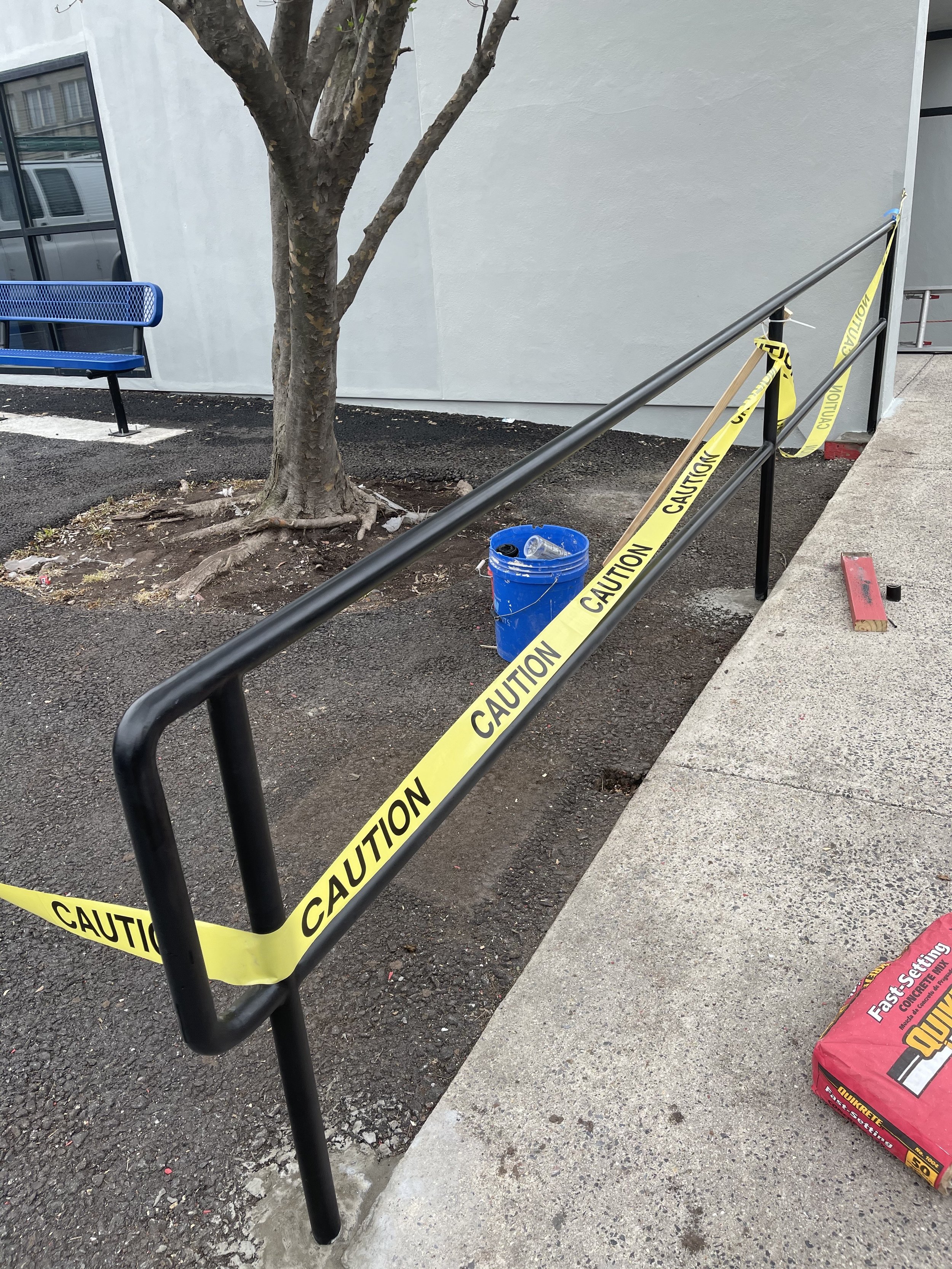 A black metal handrail with yellow caution tape wrapped around it surrounding a small dirt area with a tree located on a business ramp. There is a blue trash can with trash inside it and a red bag of quickrete on the sidewalk.