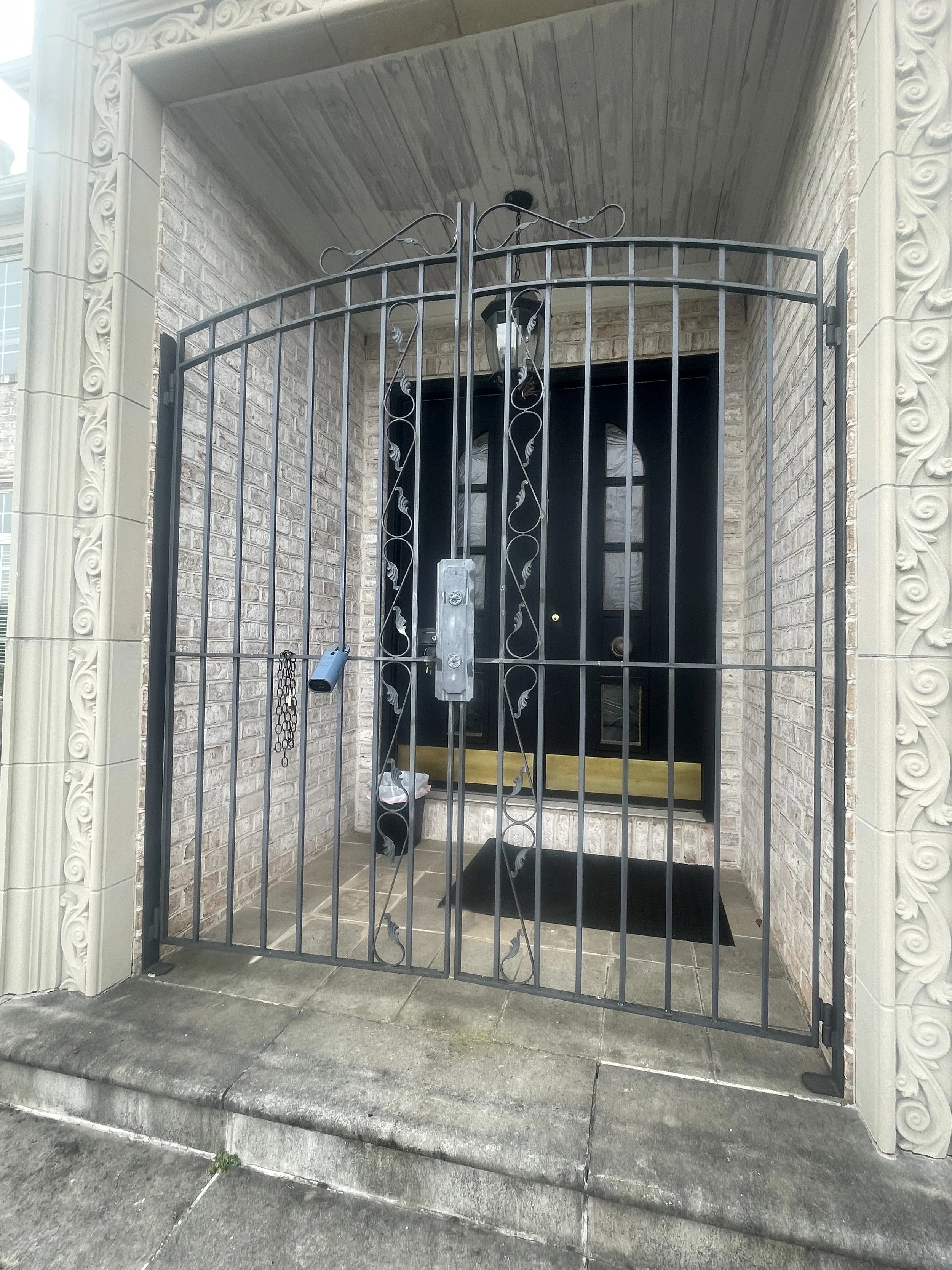 Black metal security gate at the entrance of a house, with an ornate design on the sides, leading to a black front door with arched windows. A padlock hangs on the gate.