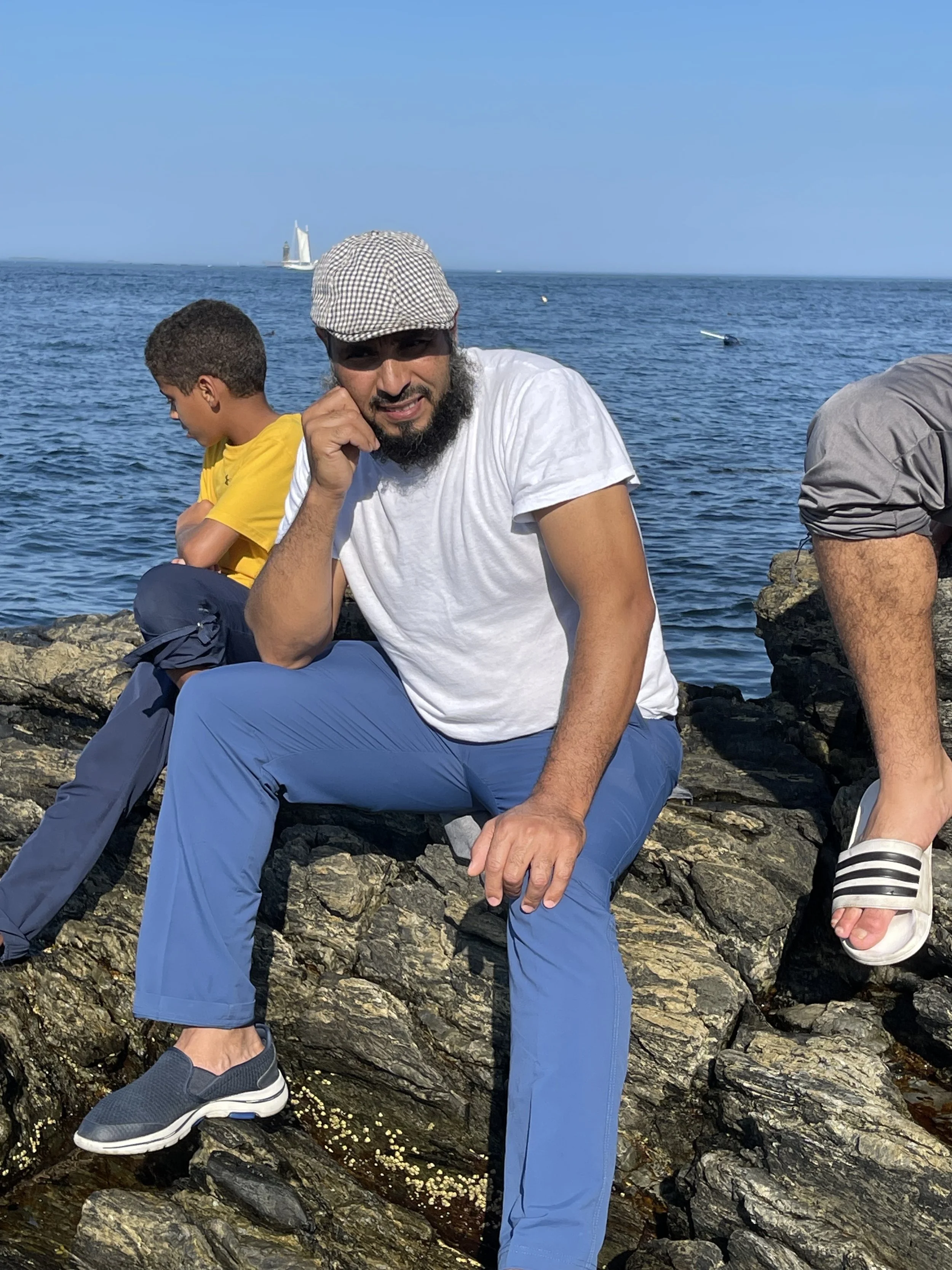 Man with a beard wearing a white t-shirt, blue pants, gray sneakers, and a checkered cap sitting on rocks near the ocean with two children, one in a yellow shirt and another in gray slide sandals, against a blue sky with sailboats in the background.