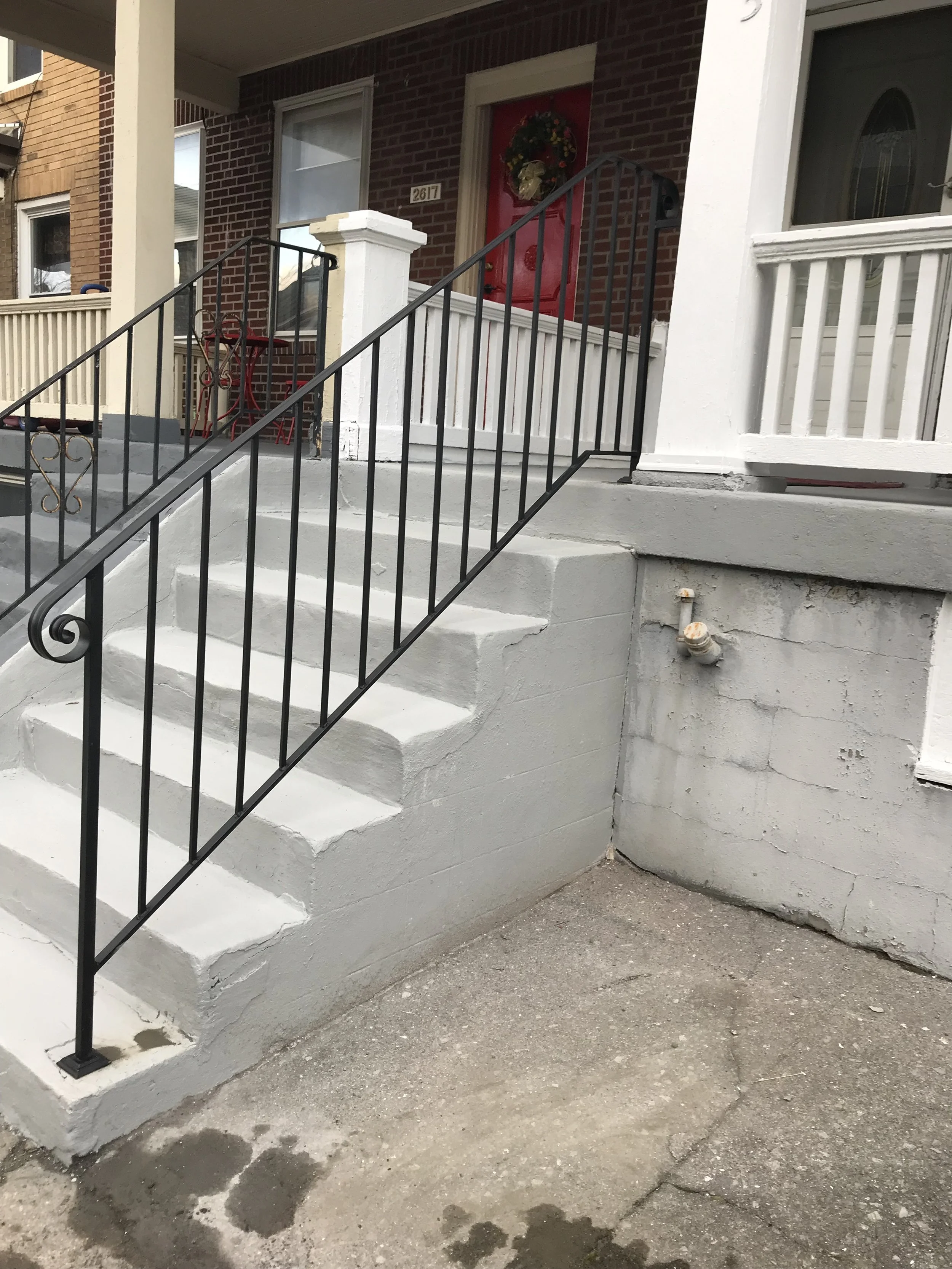 Front porch with painted concrete stairs, black metal railing, white trim, red door with holiday wreath, and brick house