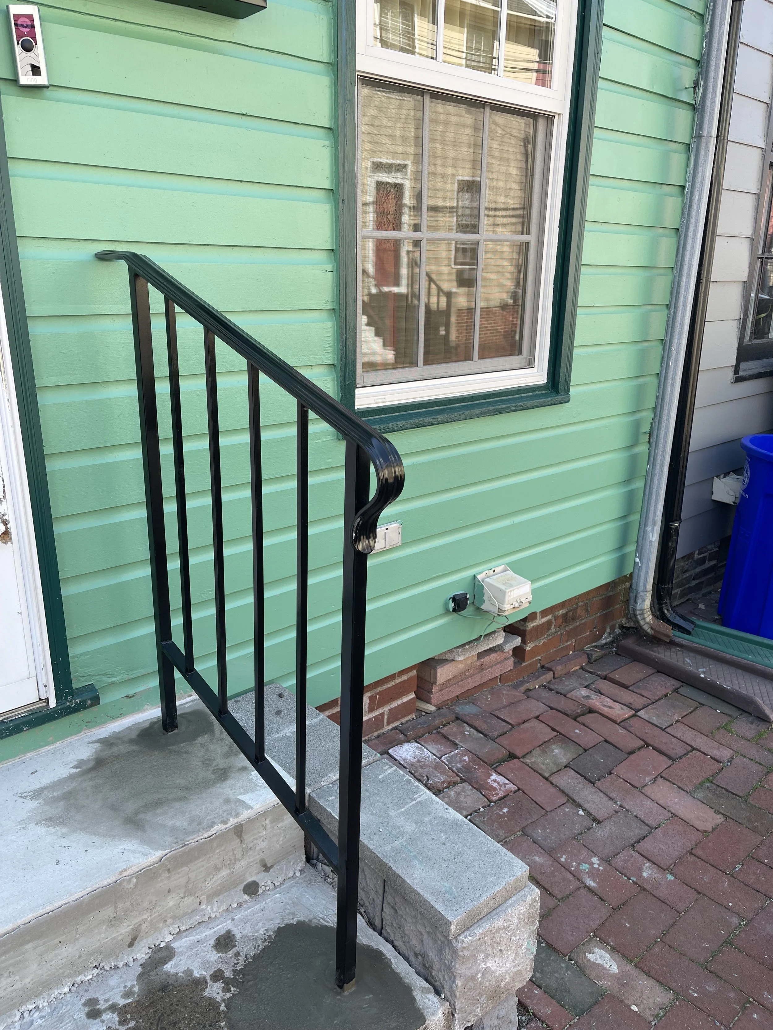 Exterior of a house with mint green siding, a window, black metal handrail, and a small concrete porch. The porch has a new layer of concrete, and there's a brick step leading up to it. To the right, there's a blue recycling bin and a metal downspout.