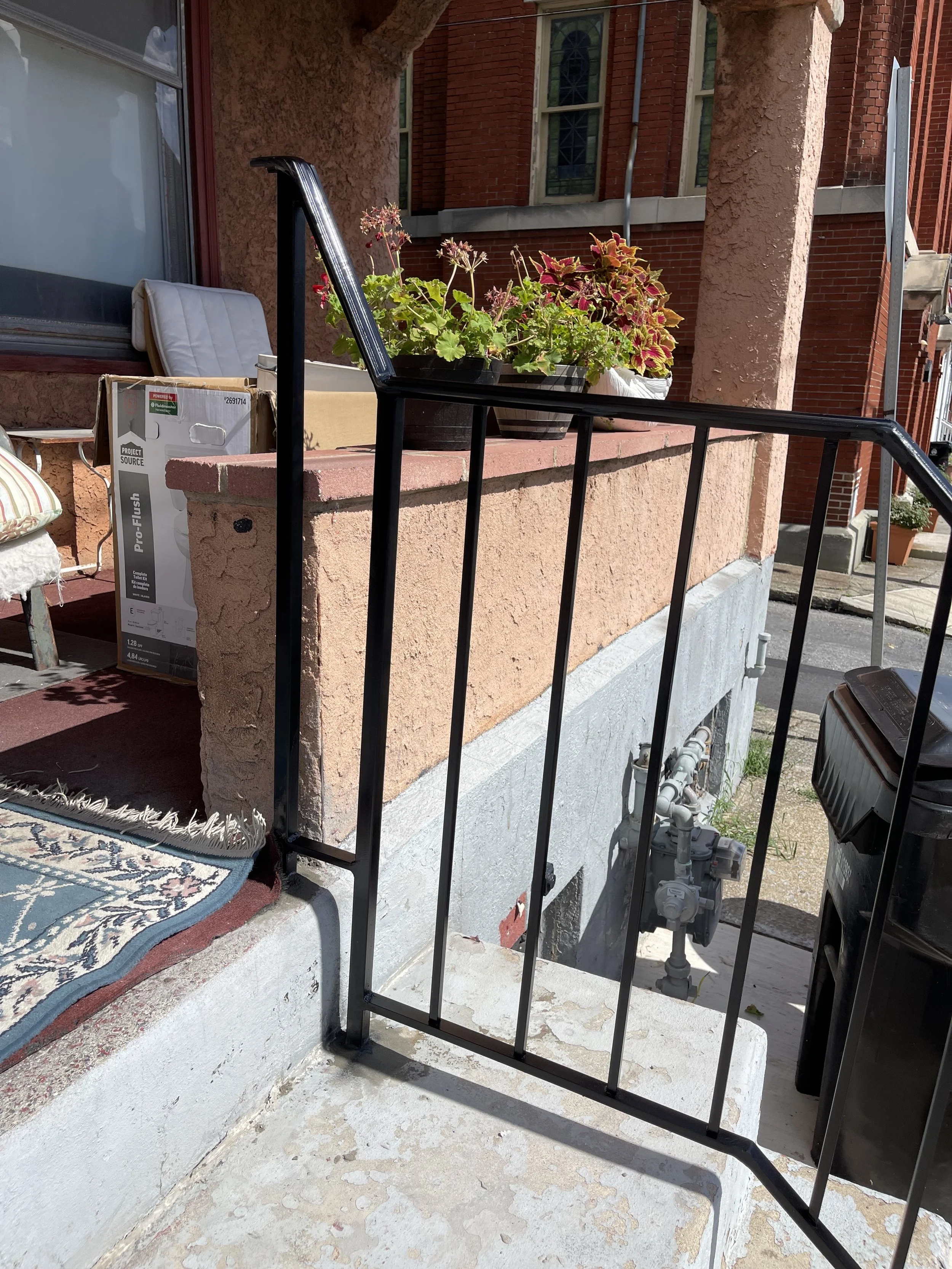 A small black metal railing at the edge of a porch, with a potted plant with pink and green leaves placed on the ledge. There is a cardboard box, a chair with a striped cushion, and a door or window with a glass pane in the background. The house has red brick walls and a stone foundation.