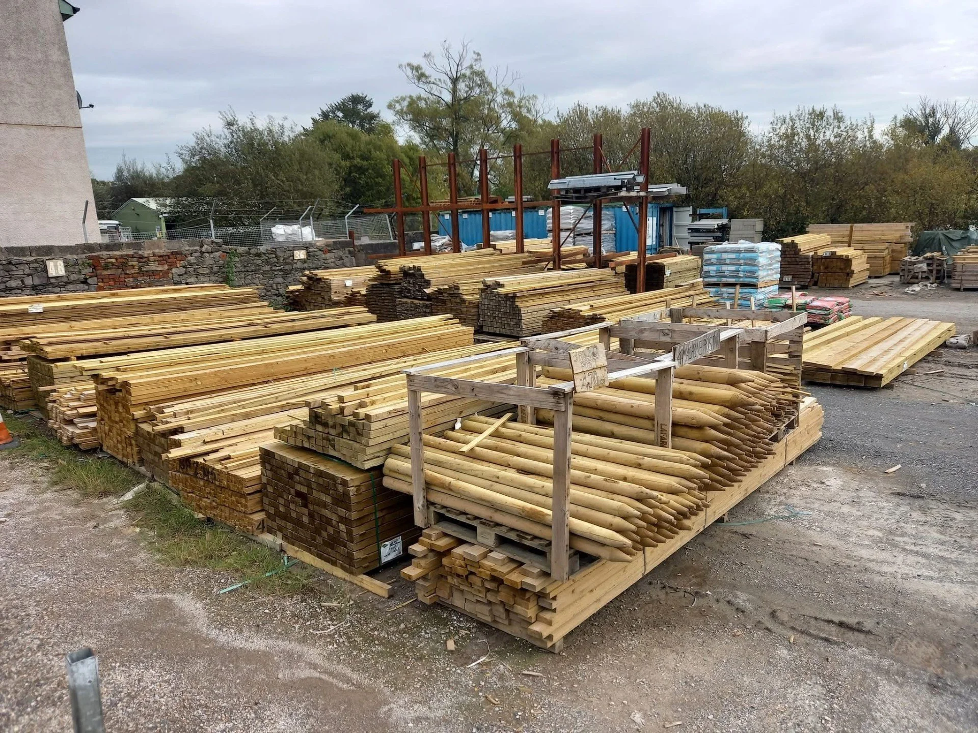Outdoor lumber yard with stacks of cut wood and wooden frames, gravel ground, trees in the background, and overcast sky.