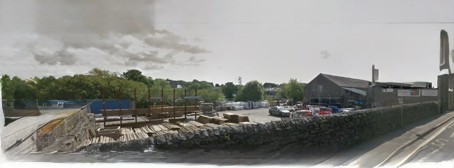 View of a construction yard with wooden logs and building materials, partly enclosed by a stone wall, with a parking lot and a building in the background, on a cloudy day.